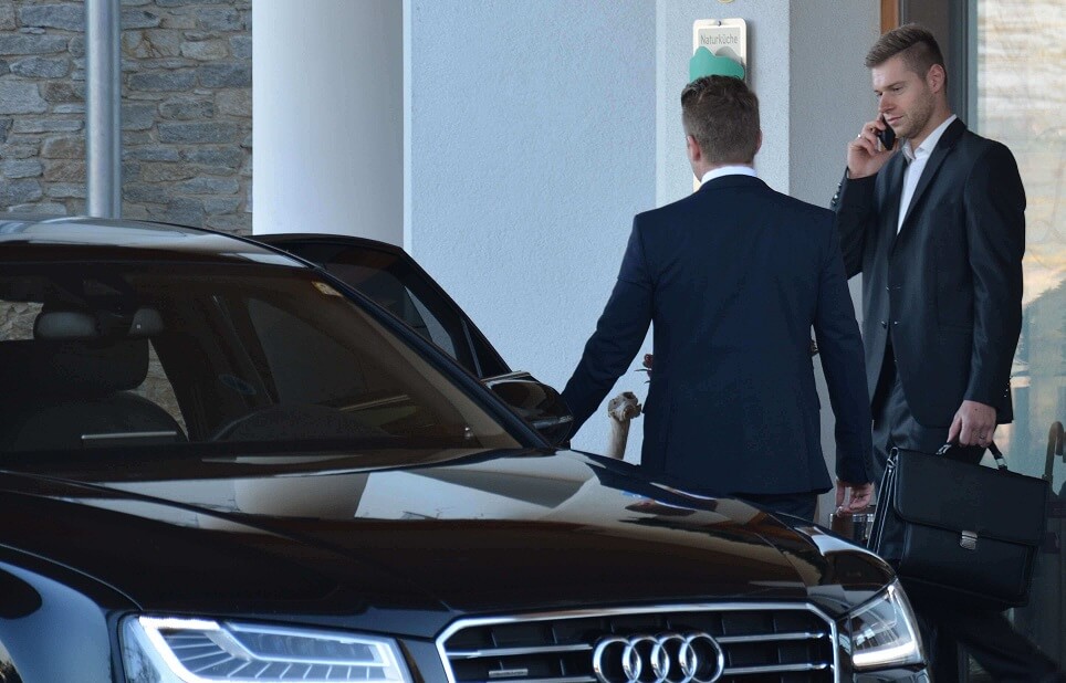Geschäftsmänner in Anzügen vor einem schwarzen Audi, einer telefoniert, der andere hält eine Aktentasche.