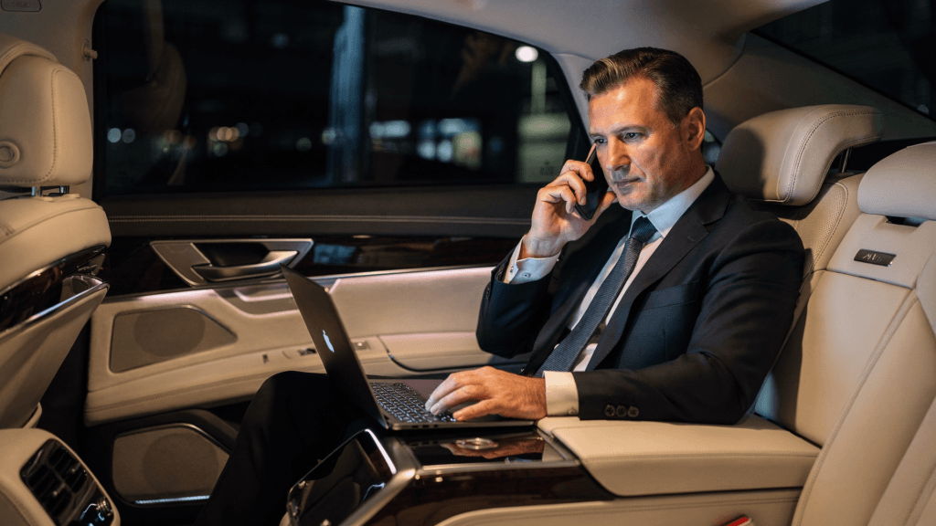 Geschäftsmann im Anzug telefoniert und arbeitet am Laptop auf dem Rücksitz eines Autos bei Nacht.
